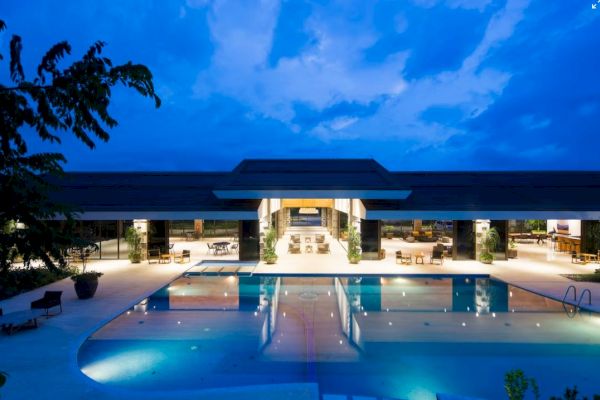An illuminated luxurious building with large glass windows and an inviting outdoor pool, set against a twilight sky.