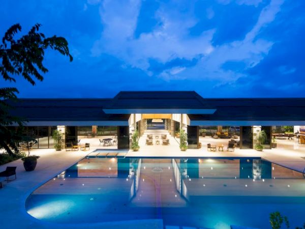 An illuminated luxurious building with large glass windows and an inviting outdoor pool, set against a twilight sky.