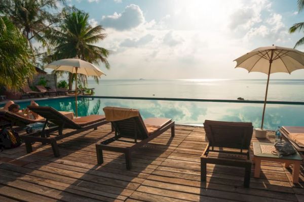 The image shows a serene poolside area with lounge chairs, umbrellas, and a breathtaking view of the ocean under a partly cloudy sky.
