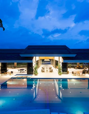 A modern building with an illuminated pool at dusk, featuring open seating areas and outdoor lighting under a partially cloudy sky.