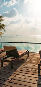 A serene poolside setup with lounge chairs, umbrellas, palm trees, and a view of a calm ocean under a partly cloudy sky at sunset.