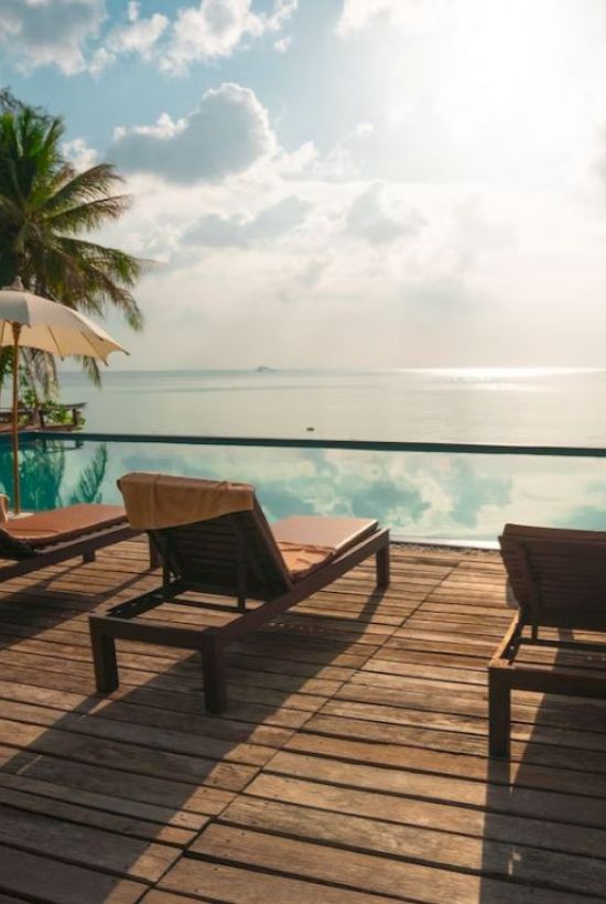 A serene tropical setting with lounge chairs, umbrellas, and a pool overlooking a calm ocean under a bright sky with scattered clouds, ending the sentence.