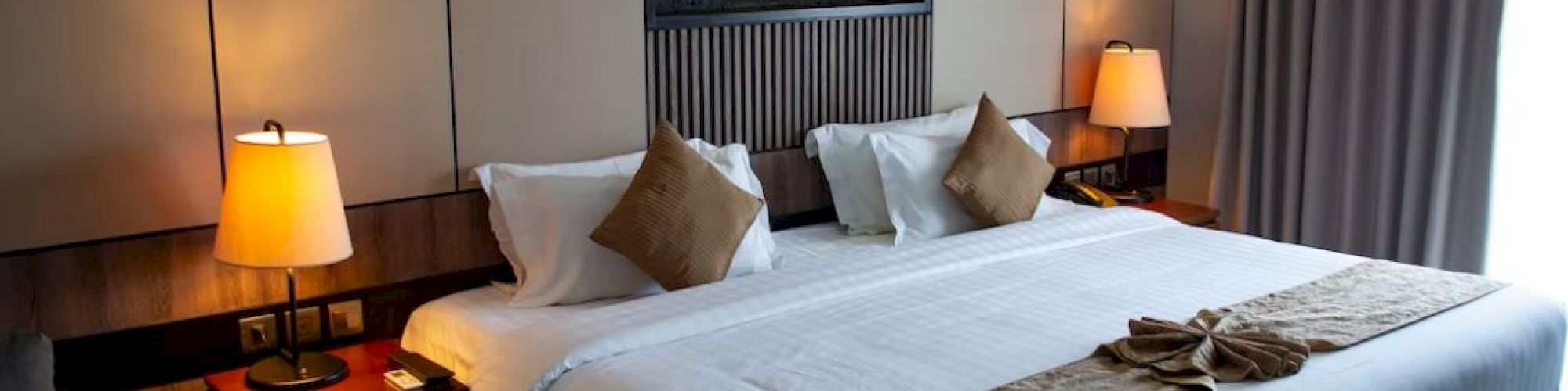 The image shows a neatly arranged modern hotel room with a large bed, bedside tables with lamps, decorative pillows, and a framed wall art piece.