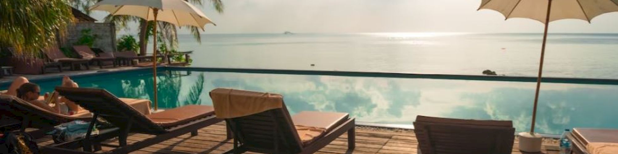 A serene tropical poolside scene with wooden lounge chairs, umbrellas, palm trees, and a beautiful view of the ocean and sky.
