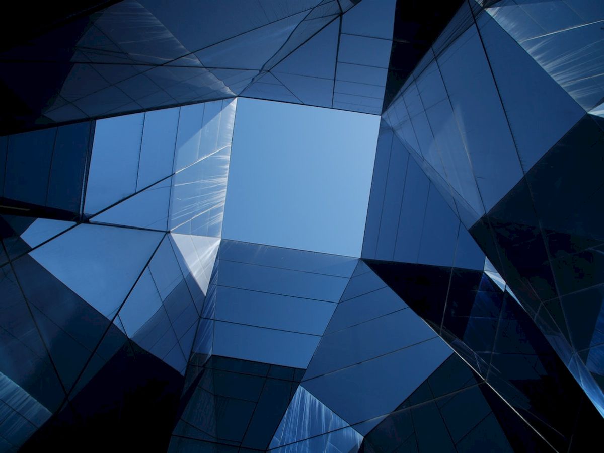 The image shows a view looking up through intersecting blue glass buildings, creating a geometric pattern with the sky visible in the center.