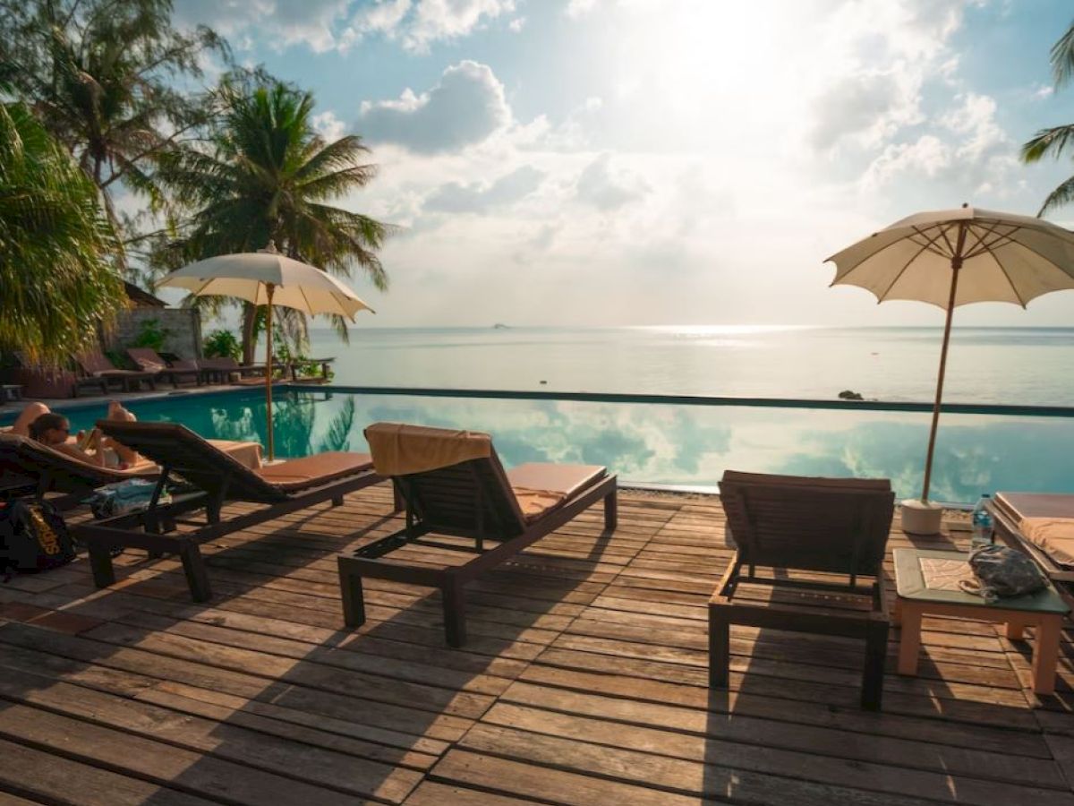 A serene poolside scene with lounge chairs, umbrellas, palm trees, and a tranquil sea view under a partly cloudy sky during sunset.