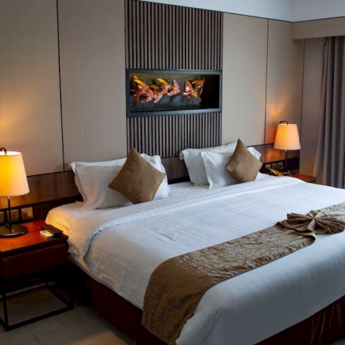 A modern hotel room with a large bed, two bedside tables with lamps, a decorative painting above the bed, and a window with curtains.