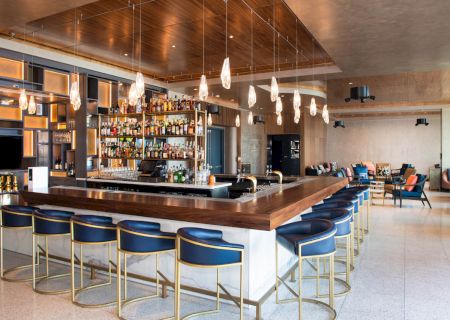 A modern bar with blue bar stools, a stocked back shelf, hanging lights, and lounge seating in a spacious, well-lit environment.