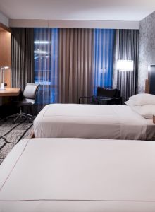 The image shows a modern hotel room with two double beds, a flat-screen TV, a desk with a chair, and warm lighting.
