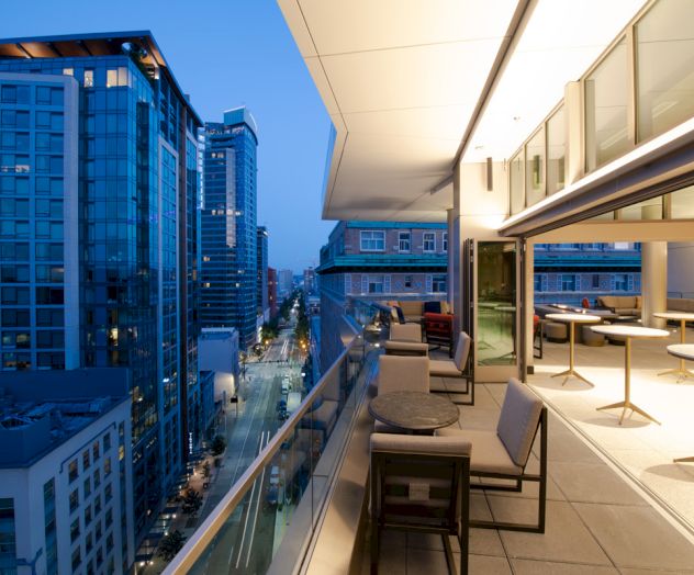 A modern cityscape view from a stylish balcony at dusk, with seating areas and tall buildings lining the street.