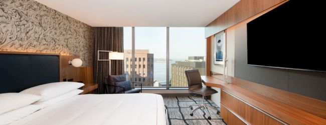 A modern hotel room with a large bed, work desk, chair, wall-mounted TV, and floor-to-ceiling windows with a cityscape view in the background.