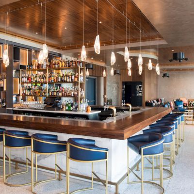 The image shows a modern bar with elegant lighting, a well-stocked counter, blue bar stools, and a lounge seating area in the background.
