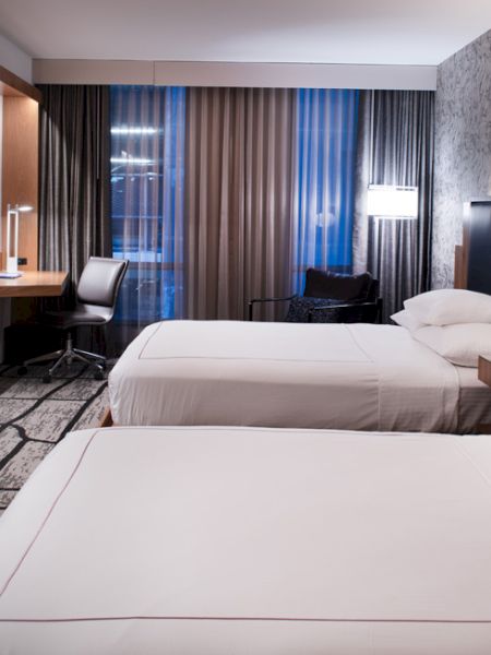 The image shows a modern hotel room with two neatly made beds, a work desk, a large wall-mounted TV, and stylish decor, including wall patterns and lighting.