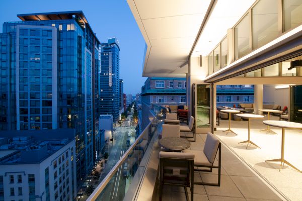 A modern balcony overlooks a cityscape with high-rise buildings. The balcony is furnished with tables and chairs, extending into an indoor area.