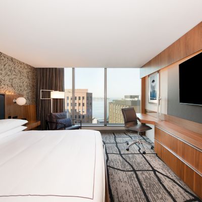 A modern hotel room with a large bed, wall-mounted TV, desk, chair, and window with a city view. The decor is contemporary and stylish.