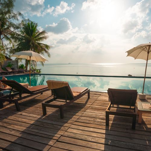 Sun loungers and umbrellas by a poolside, overlooking a serene beach with calm waters and palm trees, under a bright, partly cloudy sky.
