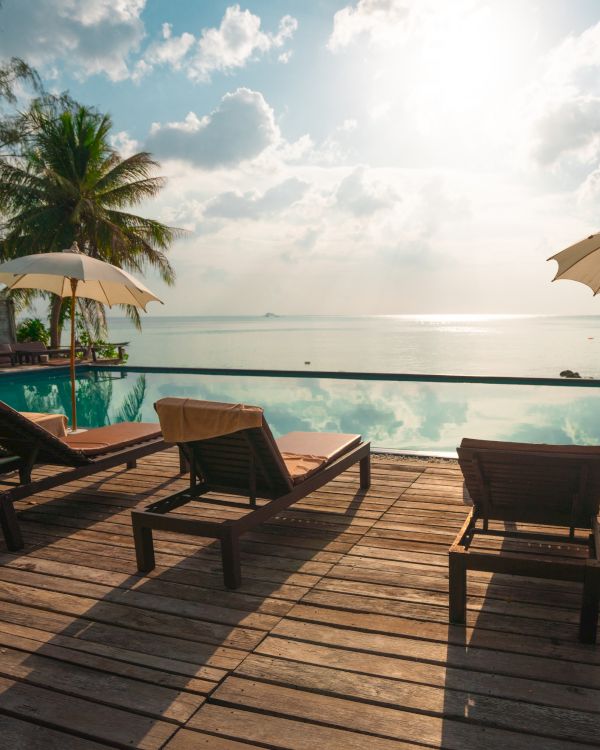 Sun loungers and umbrellas by a poolside, overlooking a serene beach with calm waters and palm trees, under a bright, partly cloudy sky.