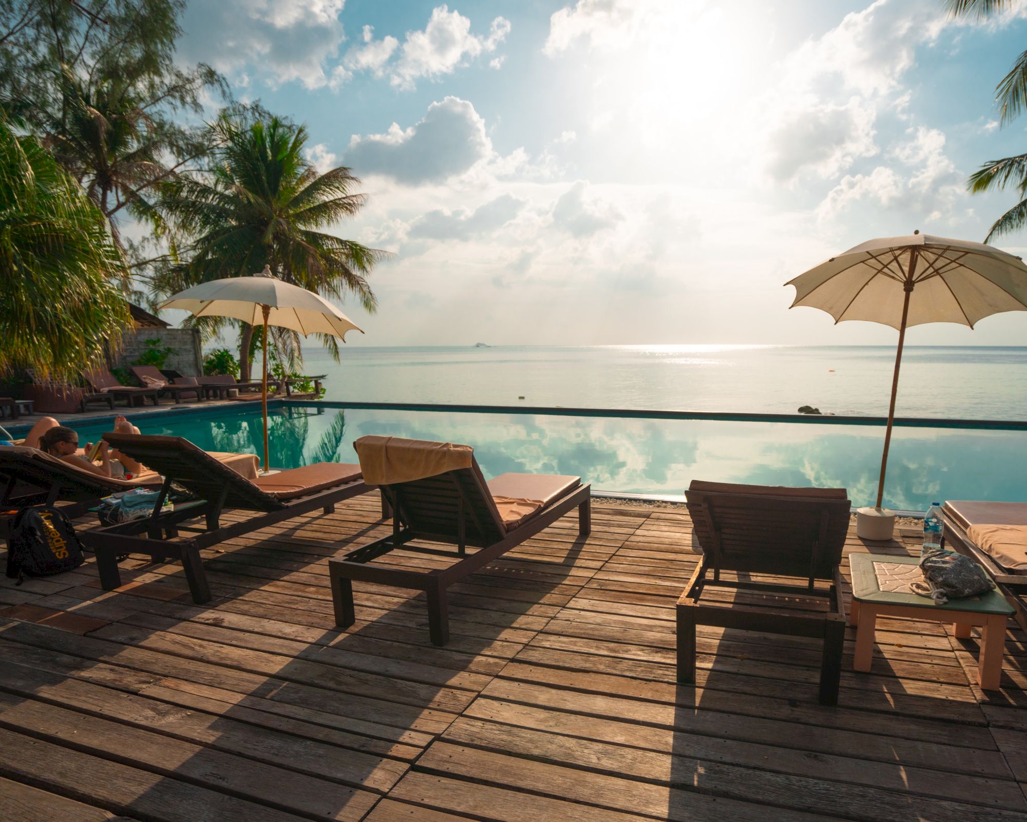 A serene poolside scene with lounge chairs, umbrellas, and a view of the ocean, surrounded by palm trees under a partly cloudy sky.
