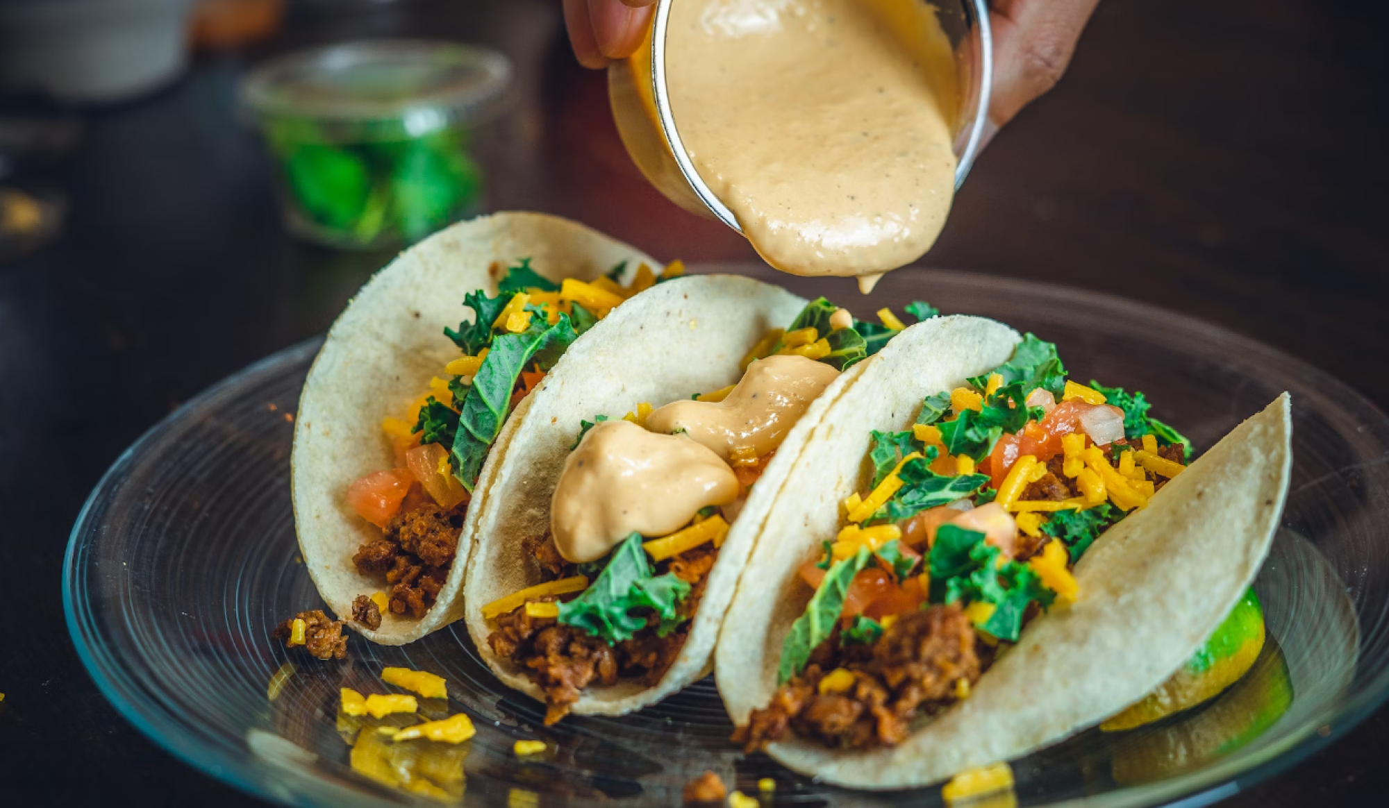 Three tacos filled with ground meat, cheese, lettuce, and tomatoes, being drizzled with a creamy sauce from a small cup on a glass plate.