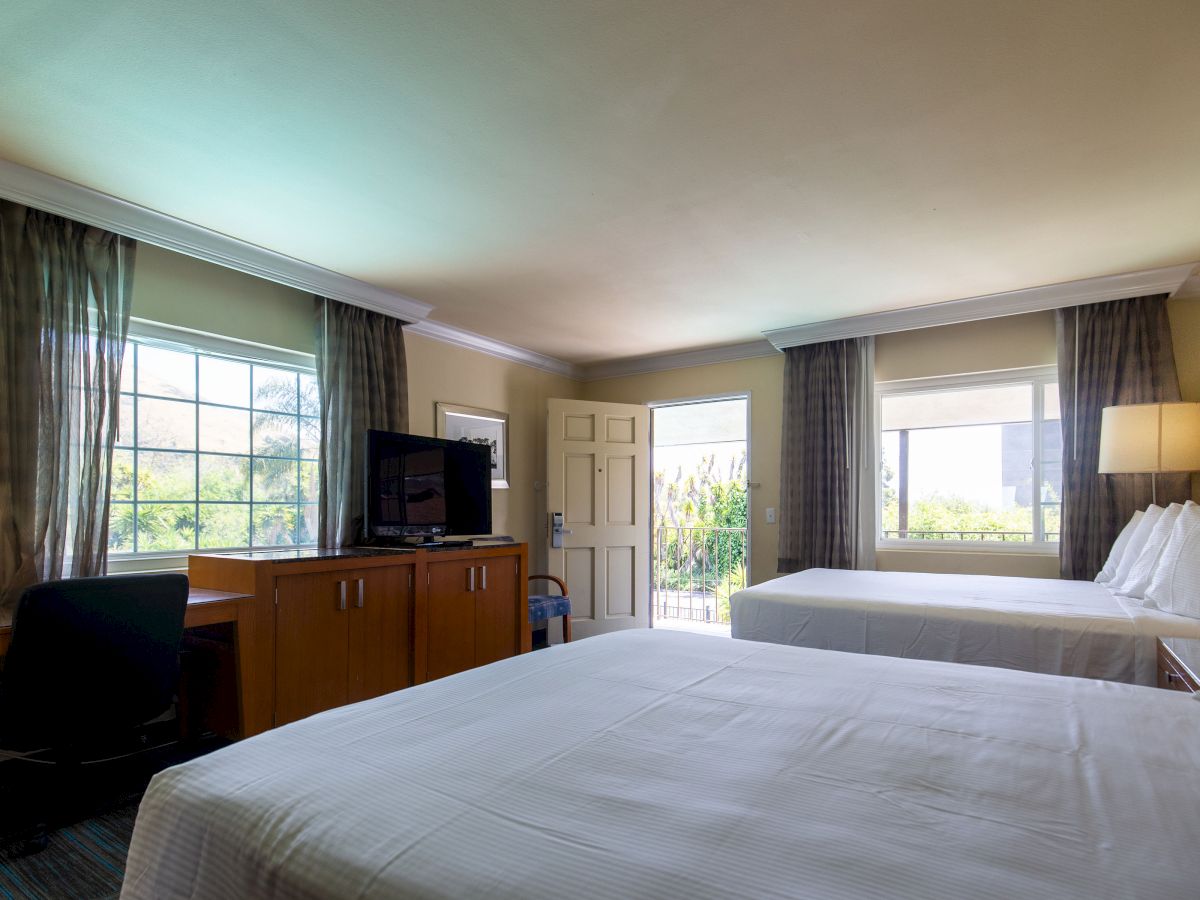 This image shows a well-lit hotel room with two beds, a TV, a desk, and a door leading outside. The room is modern with neutral decor.