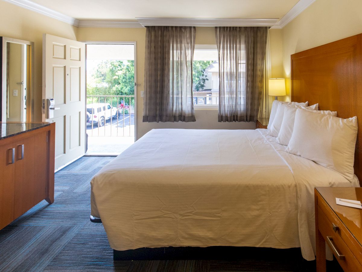 A well-lit hotel room with a neatly made bed, two nightstands with lamps, a dresser, an open door, and windows with sheer curtains.