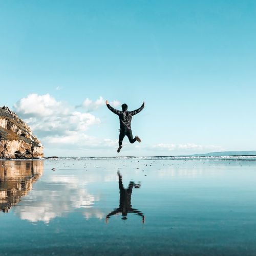 A person is jumping on a serene beach with their reflection visible on the wet sand, with cliffs on the left and a clear sky above.