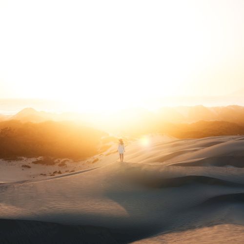 A person walks alone on sand dunes, with a bright sun setting or rising in the background, creating a serene and warm atmosphere.