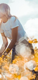 A young woman stands amidst a field of yellow flowers, with bokeh effects enhancing the sunny, dreamy atmosphere.