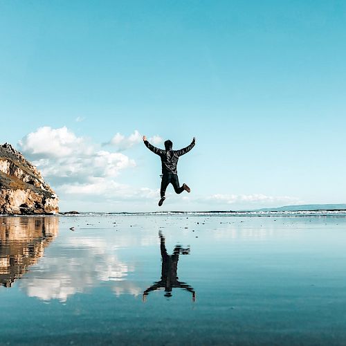 A person is jumping with arms raised on a beach, reflected perfectly in the shallow water, with a clear blue sky and rocky cliffs in the background.