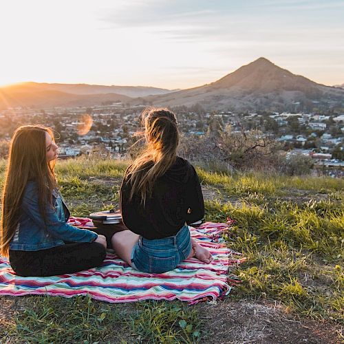 Two people sitting on a blanket atop a hill, watching the sunset over a distant town and mountains, enjoying a peaceful moment together.