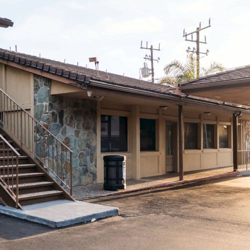 The image shows the exterior of a modest building or motel, with a staircase on the left, several windows, and a trash bin in front.