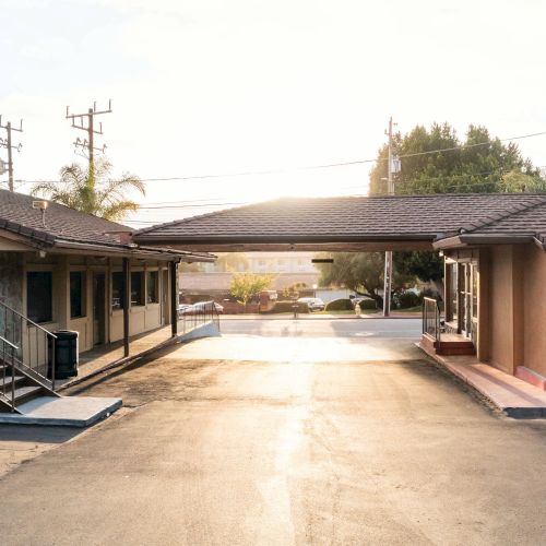 The image shows a small motel with two buildings, a parking area, and a metal staircase to the left. The sun is setting in the background.