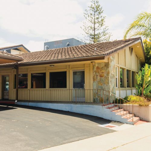 A one-story building with a sloped roof, stone facade, and several windows, surrounded by palm trees and greenery, with a paved driveway in front.