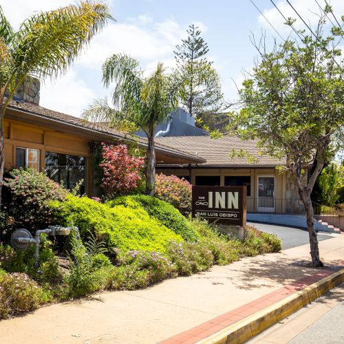 The image shows a quaint street with lush greenery and an inn sign. The sidewalk is lined with trees, and the sky is clear and bright.