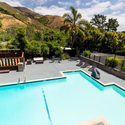 A swimming pool with lounge chairs, surrounded by lush greenery and mountains in the background, under a partly cloudy sky.