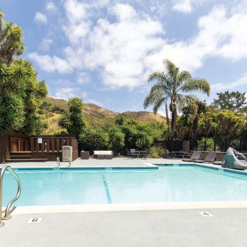 A serene outdoor pool area with lounge chairs, palm trees, and mountains in the background under a partly cloudy sky.