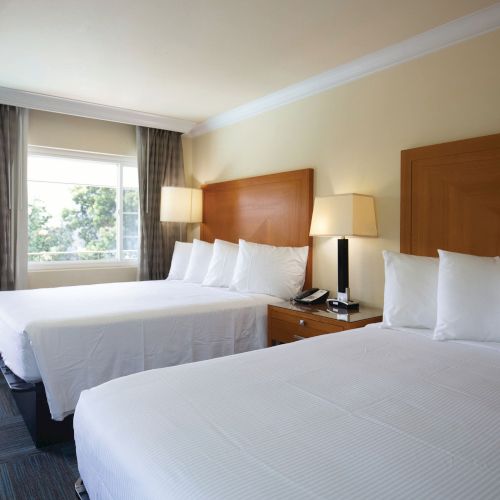 The image shows a hotel room with two neatly made double beds, bedside tables with lamps, a window with curtains, and a door.