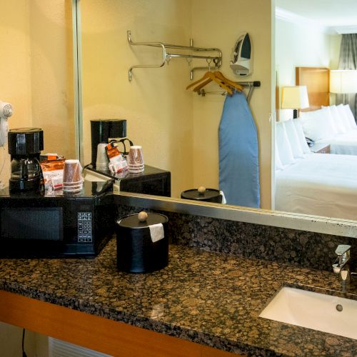 A hotel room features a vanity with a coffee maker, microwave, hairdryer, iron, ironing board, and a view of a bedroom with two beds in the reflection.