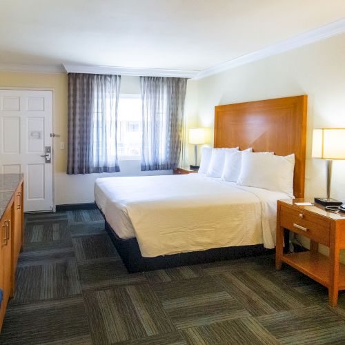 A hotel room with a neatly made bed, two side lamps, a TV on a wooden cabinet, a work desk, a chair, and a window with curtains.