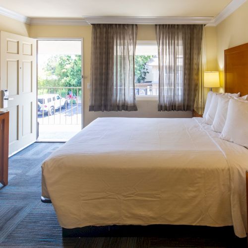 A well-lit hotel room with a large bed, wooden furniture, an open door showing a balcony, and two windows with sheer curtains.