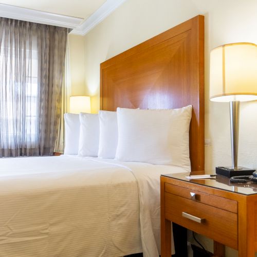 The image shows a tidy hotel room with a neatly made bed, bedside table, telephone, and two lamps. Sunlight enters through a window with curtains.