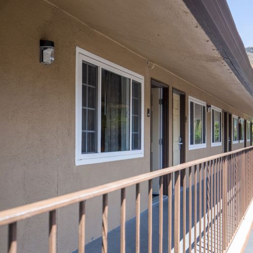 The image shows a second-floor balcony hallway of a beige building with windows and doors. A mountainous landscape is visible in the background.