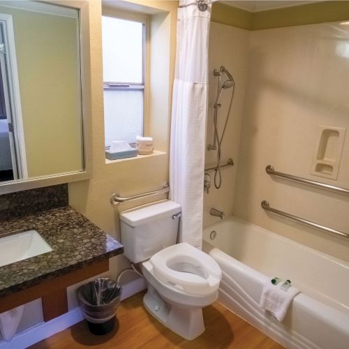 This image shows a bathroom with a sink, a toilet, and a bathtub equipped with grab bars. There is a mirror and some towels, with a glimpse of a bedroom.