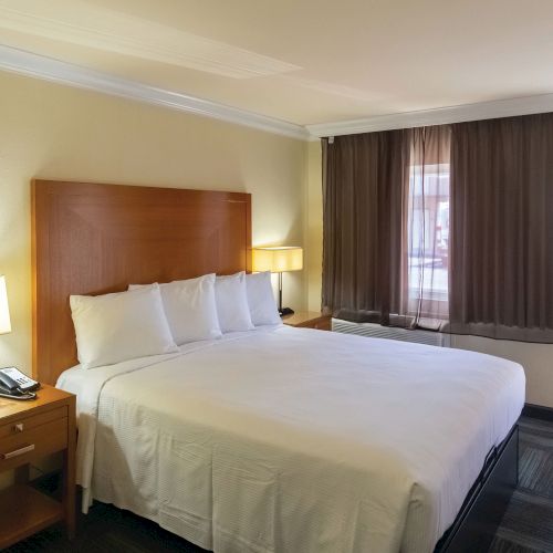 A hotel room with a king-sized bed, white linens, bedside tables, lamps, and a window with curtains. The room has neutral-colored walls.