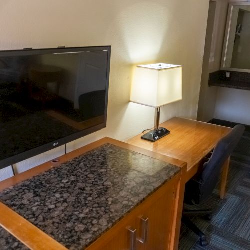 The image shows a hotel room with a TV on the wall, a desk with a chair, a lamp, and a granite top cabinet against a wall.