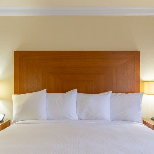 A neatly made bed with four white pillows, flanked by two nightstands with lamps. There are phones and an alarm clock on the nightstands.