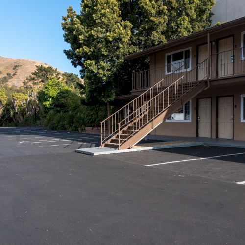 The image shows an empty parking lot adjacent to a two-story building with outdoor stairs and surrounded by trees and hills in the background.