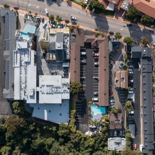 Aerial view of a residential/commercial area with buildings, parking lots, and two swimming pools, surrounded by roads and greenery.