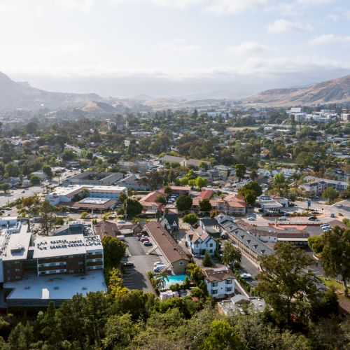 Aerial view of a city area with scattered buildings, greenery, and surrounding hills in the background under a partly cloudy sky.