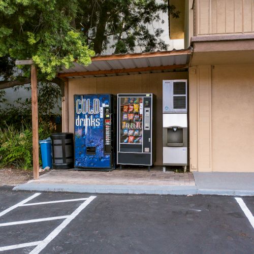 A vending machine area with a soda machine, a snack machine, and an ice dispenser next to a beige building.